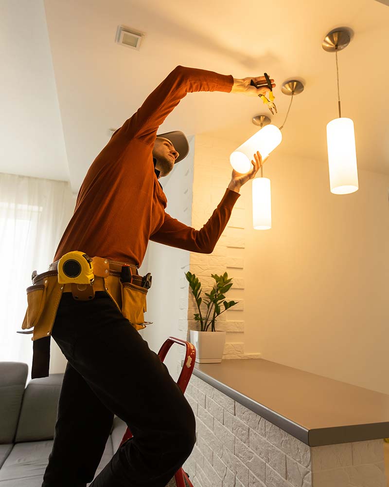 Electrician on a ladder installing a bulb in a high ceiling fixture, a routine service for Greenfield residences.