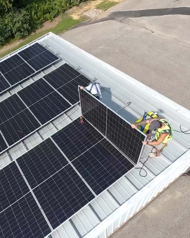 Two workers positioning a small business solar panel on a storage building roof with most of the array already installed